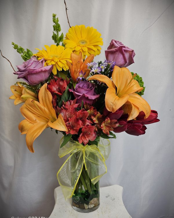A clear vase with a colorful arrangement of flowers including yellow daisies, purple roses, orange lilies, red alstroemerias, and other assorted blooms, set against a white background.