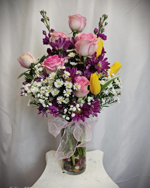 A flower arrangement in a clear glass vase with pink roses, white baby's breath, purple daisies, and yellow tulips, accented with a pink ribbon bow, placed on a small white table.