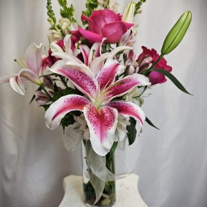 A clear vase holding a bouquet of various flowers including pink lilies, roses, and snapdragons, set against a plain white background.