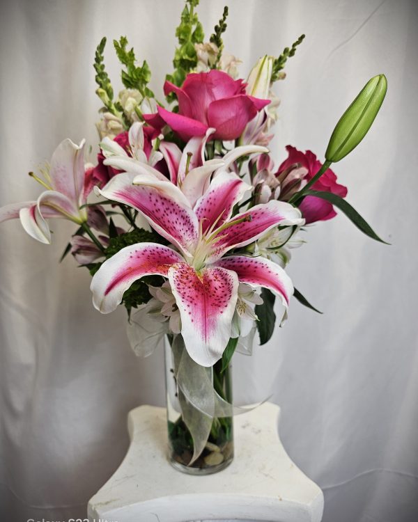 A clear vase holding a bouquet of various flowers including pink lilies, roses, and snapdragons, set against a plain white background.