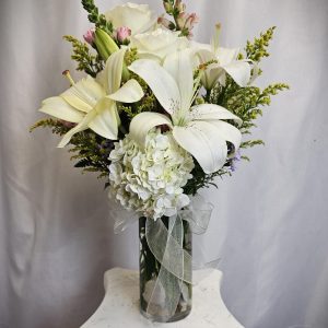 A bouquet of white flowers, including lilies and hydrangeas, arranged in a transparent vase with a white ribbon, placed on a white pedestal against a gray background.