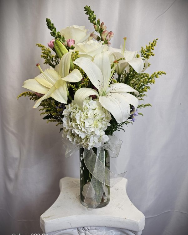 A bouquet of white flowers, including lilies and hydrangeas, arranged in a transparent vase with a white ribbon, placed on a white pedestal against a gray background.