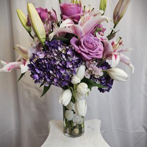 A vase holding a floral arrangement of pink lilies, purple roses, white tulips, and purple hydrangeas, placed on a white pedestal.