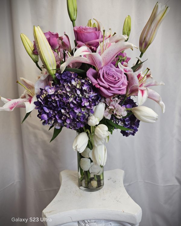 A vase holding a floral arrangement of pink lilies, purple roses, white tulips, and purple hydrangeas, placed on a white pedestal.