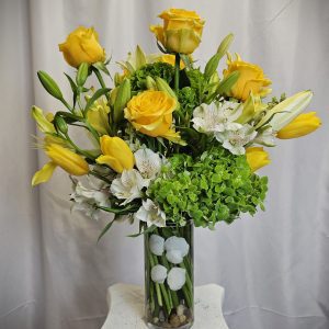 A clear vase arrangement of yellow roses, tulips, white lilies, green hydrangeas, and round white flowers, placed on a white stand against a white curtain background.