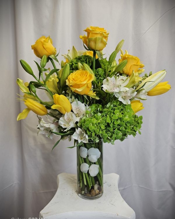 A clear vase arrangement of yellow roses, tulips, white lilies, green hydrangeas, and round white flowers, placed on a white stand against a white curtain background.