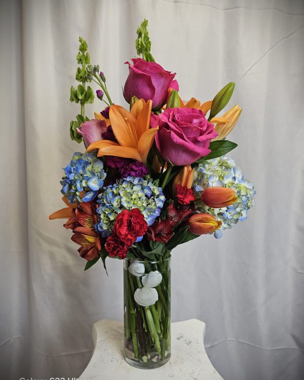A tall glass vase holds a colorful bouquet with pink roses, orange lilies, blue hydrangeas, and various other flowers, set against a plain white curtain backdrop.