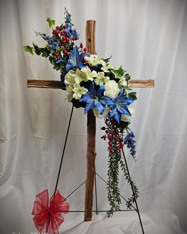A wooden cross on a stand with an arrangement of blue, white, and red flowers, and greenery draping down. A red bow is attached to the base. The background is a white curtain.