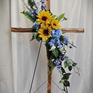 A Wooden Cross & Silk Flowers Easel, adorned with sunflowers, blue hydrangeas, and greenery, displayed on a black metal stand against a white backdrop.