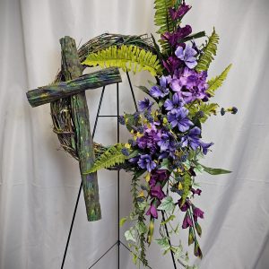 A Wooden Cross & Silk Flowers Easel with a rustic cross and vibrant purple flowers, accented with green ferns, displayed on a black easel against a white background.