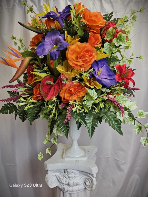 A white pedestal with a vibrant floral arrangement, featuring orange roses, purple irises, and various greenery. The photo is labeled "Wooden Cross & Silk Flowers Easel" in the bottom left corner.