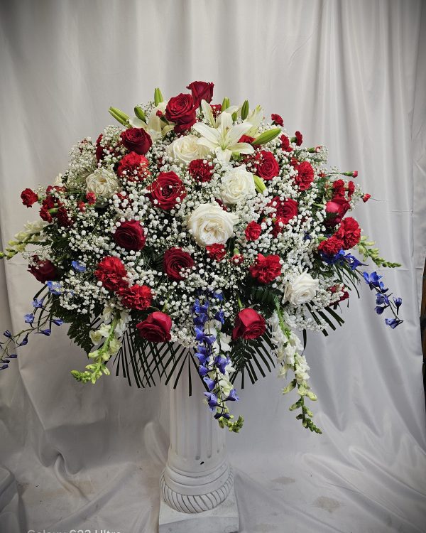 A large floral arrangement composed of red roses, white roses, baby's breath, and blue delphiniums, displayed in a tall white pedestal vase against a white background.