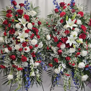 Two standing floral wreaths featuring red roses, white lilies, blue delphiniums, and baby's breath, set against a white curtain background.