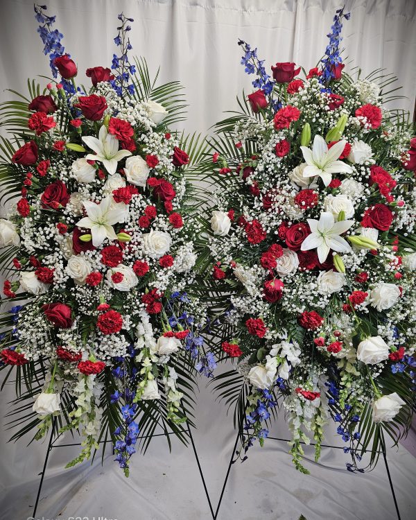 Two standing floral wreaths featuring red roses, white lilies, blue delphiniums, and baby's breath, set against a white curtain background.