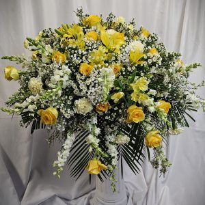 A large floral arrangement in a white vase featuring yellow roses, white lilies, baby’s breath, and assorted greenery, set against a white backdrop.