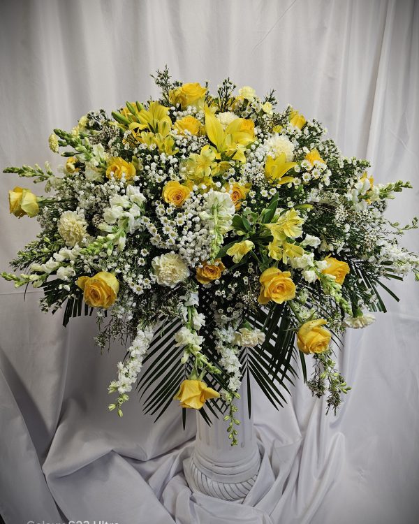 A large floral arrangement in a white vase featuring yellow roses, white lilies, baby’s breath, and assorted greenery, set against a white backdrop.