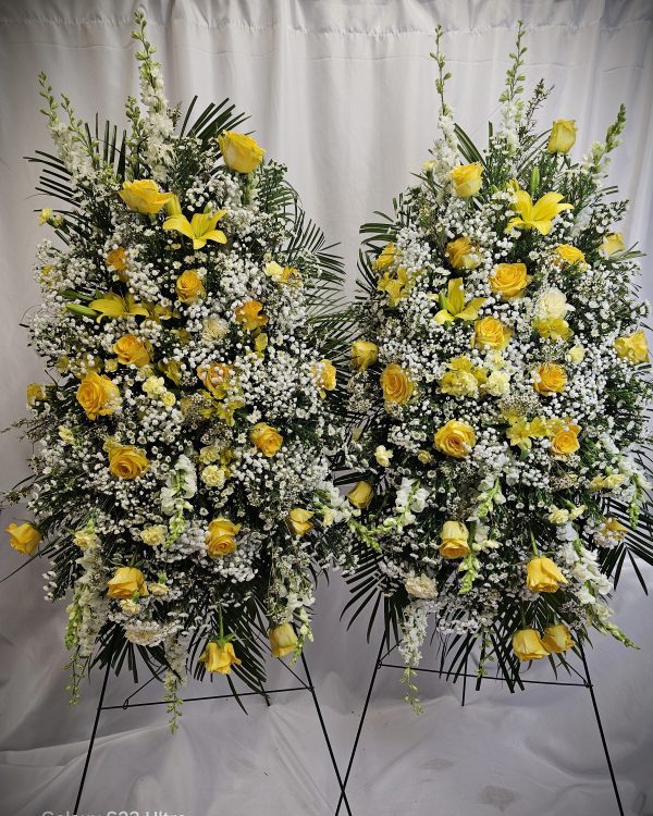 Two large floral arrangements featuring yellow roses, white baby's breath, and various green foliage, set against a white curtain background.