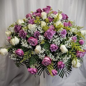 A large floral arrangement with light purple and white roses, baby's breath, yellow sprigs, and various green foliage placed in front of a white curtain backdrop.