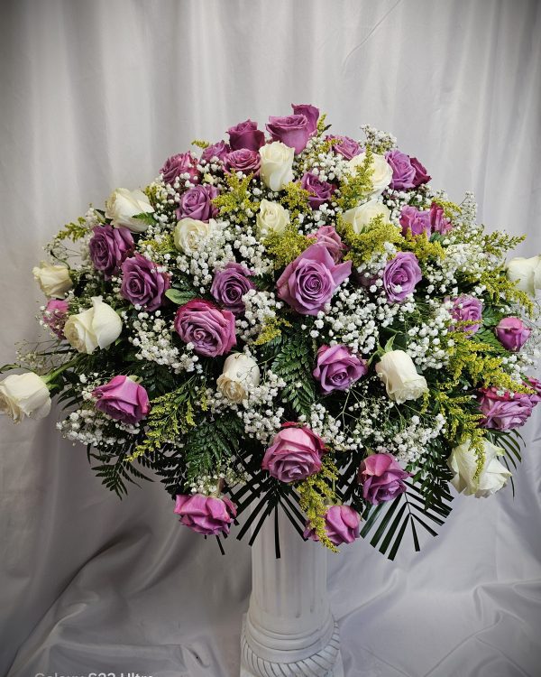 A large floral arrangement with light purple and white roses, baby's breath, yellow sprigs, and various green foliage placed in front of a white curtain backdrop.