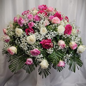 A flower arrangement in a white vase featuring pink, purple, and white roses complemented by baby's breath and green foliage, set against a white fabric backdrop.