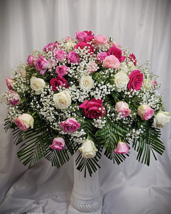 A flower arrangement in a white vase featuring pink, purple, and white roses complemented by baby's breath and green foliage, set against a white fabric backdrop.