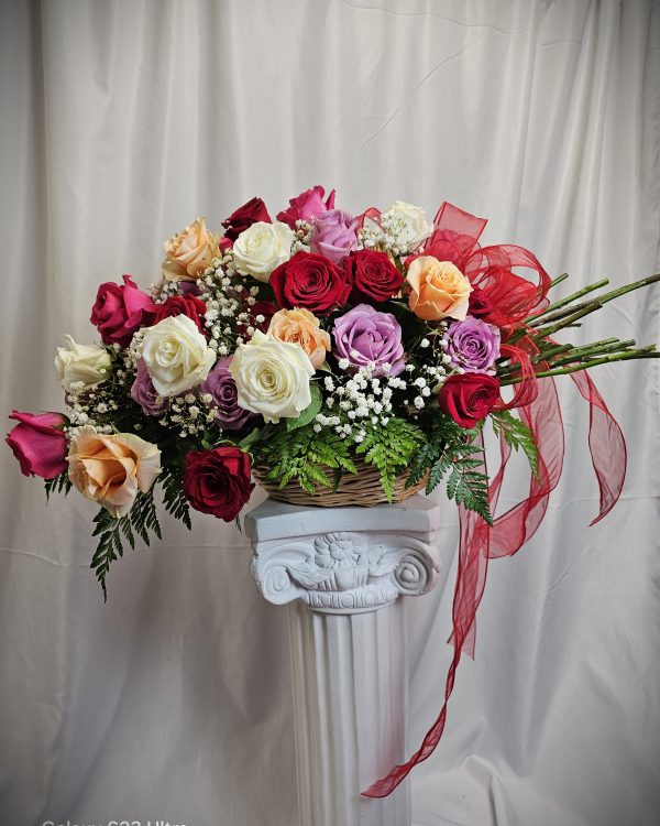 A floral arrangement with red, white, pink, and purple roses, baby's breath, and ferns in a wicker basket displayed on a white decorative pedestal. A red ribbon accents the bouquet.