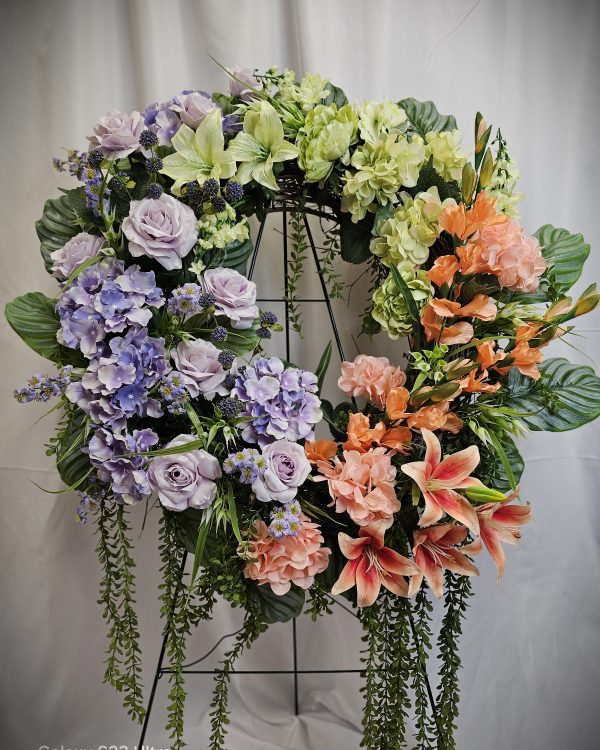 A floral wreath with a mix of purple, orange, and green flowers, including roses, hydrangeas, lilies, and greenery, displayed against a white background.