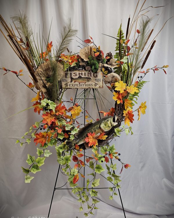 A wreath featuring a sign that reads "Fishing Expeditions" with various leaves, flowers, and a decorative fish. The wreath is mounted on a metal stand against a white background.