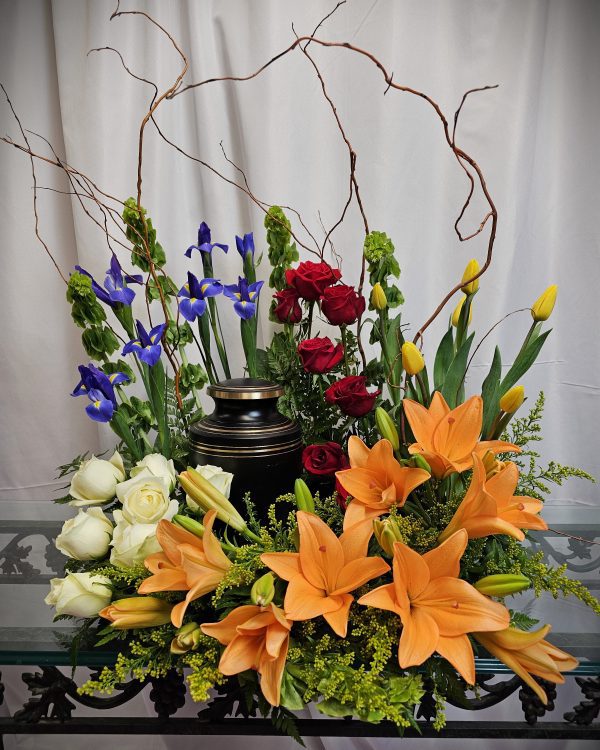 A Celebration of Life Urn Wreath surrounded by an arrangement of red roses, orange lilies, white roses, yellow tulips, and purple irises, set against a white background with twisting branches added for decoration.