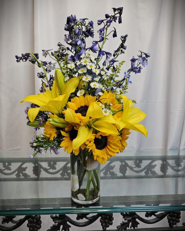 A glass vase with a bouquet of yellow lilies, sunflowers, white daisies, and purple flowers on a glass table with a white curtain as the background.