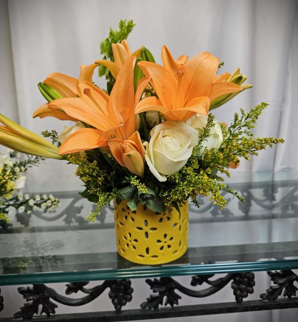 A floral arrangement featuring orange lilies and white roses in a yellow vase with a perforated design, placed on a glass shelf with a white curtain background.