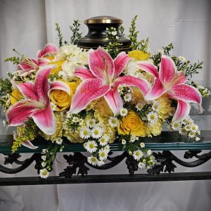 A floral arrangement featuring pink lilies, yellow roses, and white daisies is placed on a glass table. A black urn is positioned in the center of the flowers.