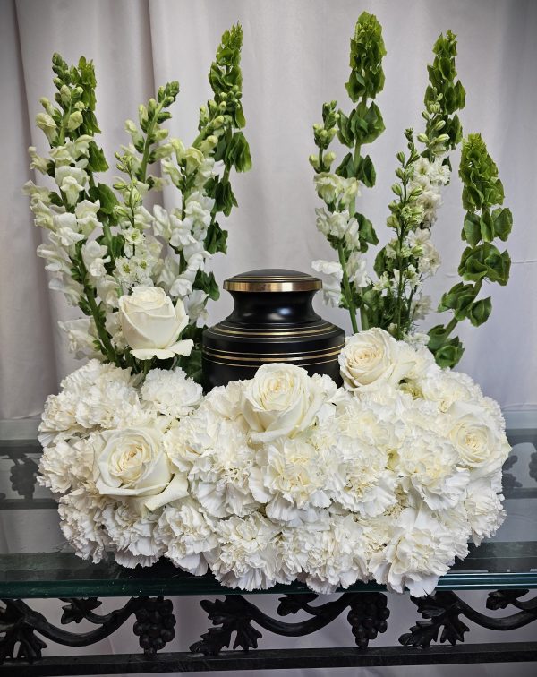 A White over White Cremation Urn Wreath is surrounded by white roses, carnations, and tall greenery on a glass table with ornate legs.