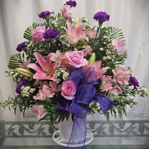 A floral arrangement with pink roses, pink lilies, purple carnations, white daisies, and green leaves in a white vase, decorated with a purple bow, placed on a glass table.