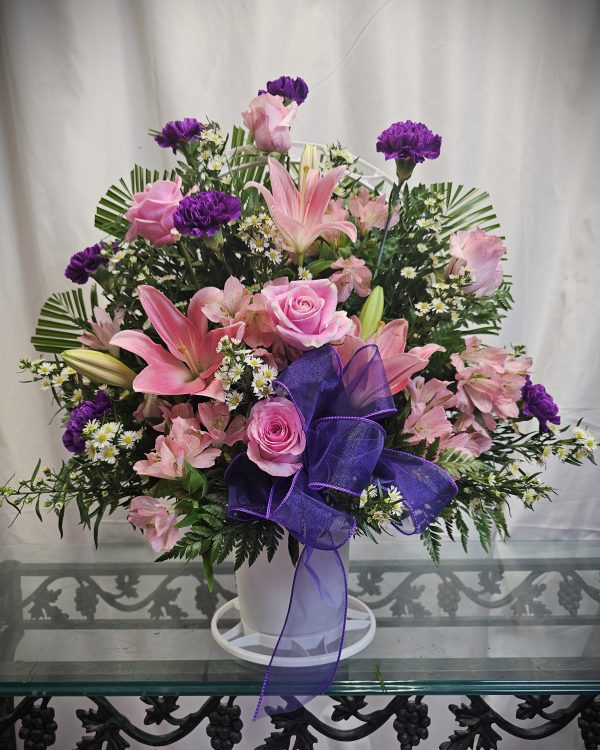 A floral arrangement with pink roses, pink lilies, purple carnations, white daisies, and green leaves in a white vase, decorated with a purple bow, placed on a glass table.