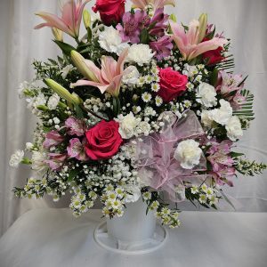 The Mix Sympathy Basket is a beautiful floral arrangement presented in a white vase, featuring an exquisite combination of pink lilies, red roses, white carnations, and baby's breath. A delicate pink ribbon is lovingly tied around the vase, all set against a plain backdrop.