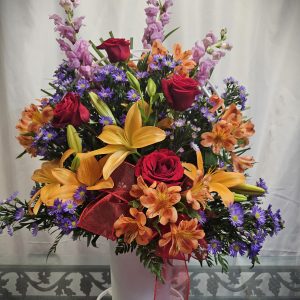 A floral arrangement with orange lilies, red roses, purple aster flowers, and pink snapdragons in a white vase with a red ribbon on a glass table against a white background.