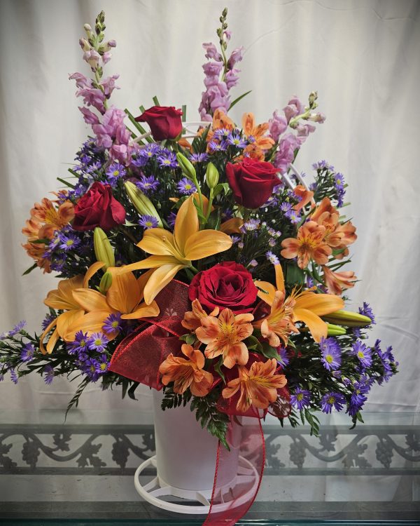 A floral arrangement with orange lilies, red roses, purple aster flowers, and pink snapdragons in a white vase with a red ribbon on a glass table against a white background.