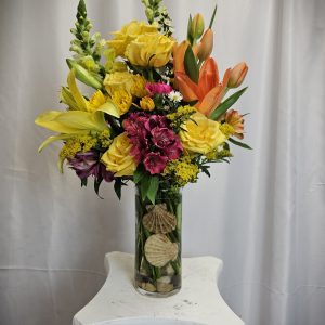 A bouquet of yellow, orange, and burgundy flowers arranged in a clear glass vase with decorative shells at the bottom, placed on a white pedestal against a light-colored curtain background.