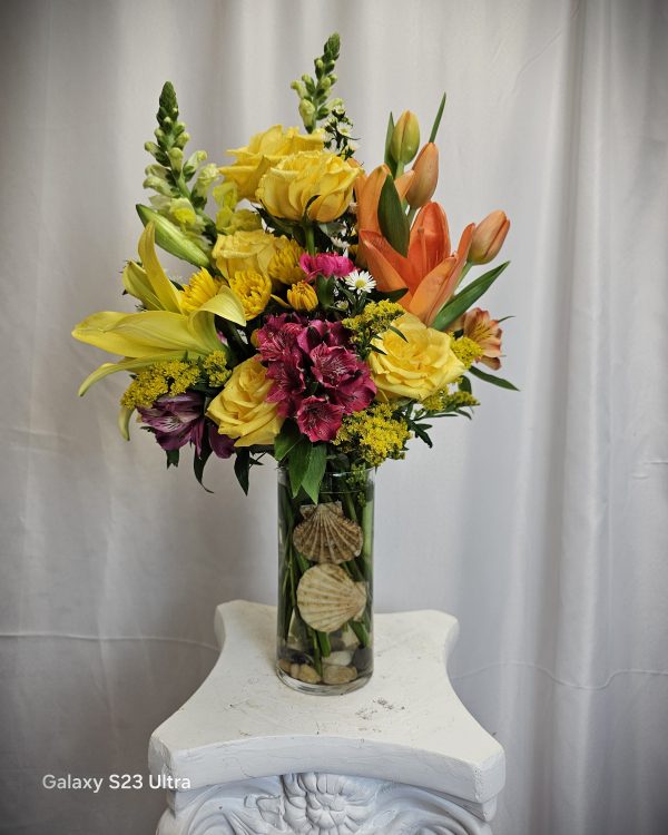 A bouquet of yellow, orange, and burgundy flowers arranged in a clear glass vase with decorative shells at the bottom, placed on a white pedestal against a light-colored curtain background.