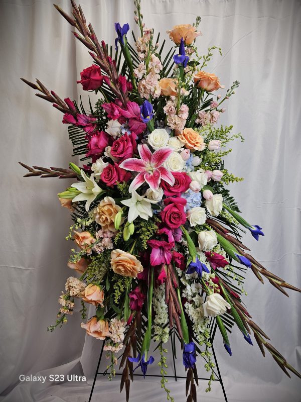 A large floral arrangement containing a variety of flowers including pink lilies, red and pink roses, white carnations, blue irises, and assorted green foliage, set against a white backdrop.