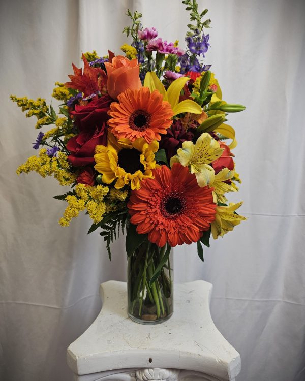 A bouquet of colorful flowers, including orange gerbera daisies, yellow lilies, and other mixed blooms arranged in a glass vase, placed on a white stool with a white curtain background.