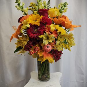 A tall glass vase filled with a vibrant arrangement of flowers, including red, yellow, and orange blooms, adorned with autumn leaves and berries, placed on a white ornate stand against a plain backdrop.