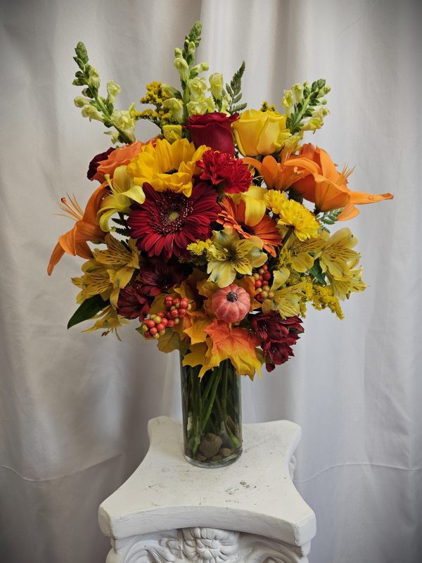 A tall glass vase filled with a vibrant arrangement of flowers, including red, yellow, and orange blooms, adorned with autumn leaves and berries, placed on a white ornate stand against a plain backdrop.