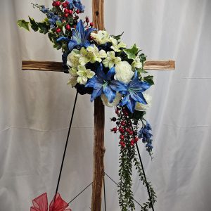 The Wooden Sympathy Cross features an arrangement of blue and white flowers, red berries, and green leaves, beautifully displayed on a stand with a red bow at the base against a white background.