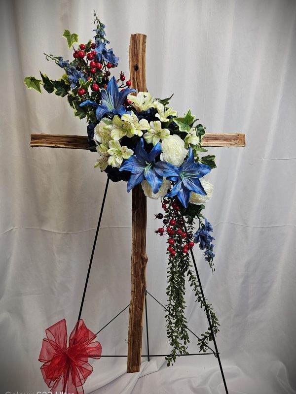 The Wooden Sympathy Cross features an arrangement of blue and white flowers, red berries, and green leaves, beautifully displayed on a stand with a red bow at the base against a white background.