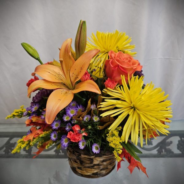 A woven basket with orange lilies, a peach rose, yellow spider mums, and other small purple and pink flowers against a white background.