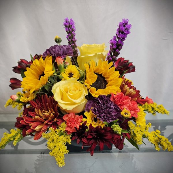 A colorful floral arrangement featuring sunflowers, yellow roses, purple liatris, red chrysanthemums, and yellow solidago on a clear glass surface.