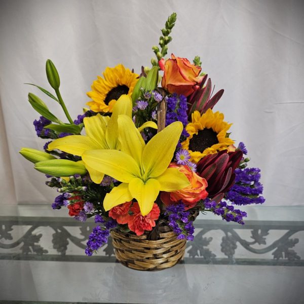 A vibrant floral arrangement in a woven basket featuring sunflowers, lilies, roses, and purple accents on a glass table with a white curtain backdrop.