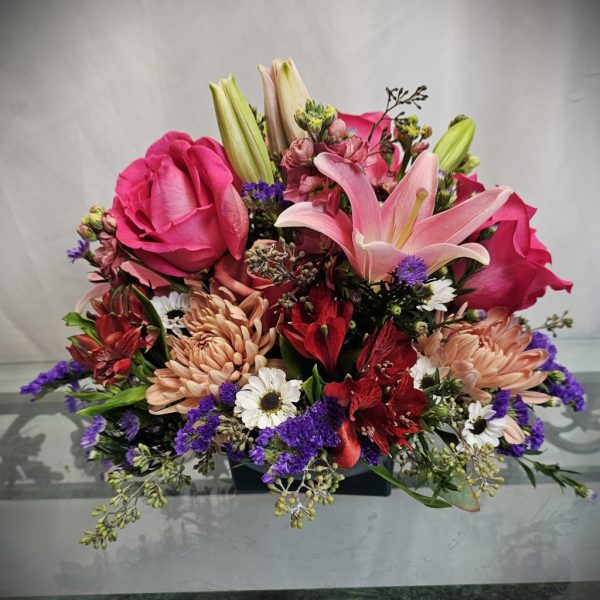 A vibrant floral arrangement featuring pink roses, lilies, red and peach flowers, purple statice, and white daisies.